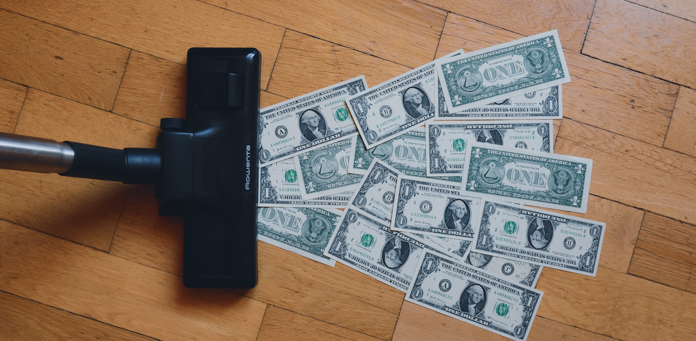 A vacuum runs over a pile of dollar bills on a hardwood floor.