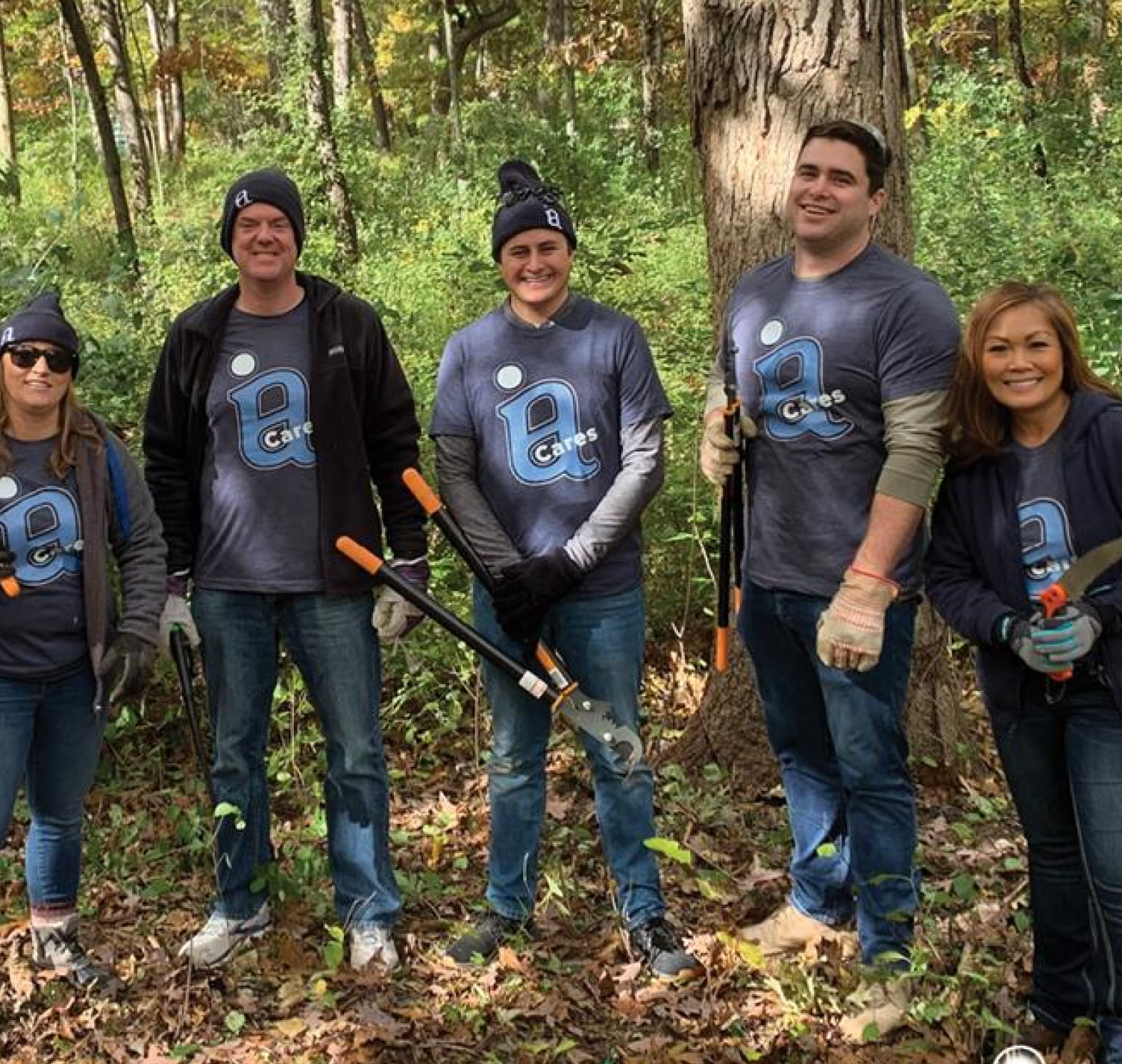 Investors Associated team participating in an IA Cares initiative. They hold tree trimmers and stand in a forest.